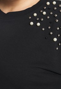 Svart tygtröja med spridda dekorativa runda pärlor i silver, svart och pärlvitt nära axelområdet.