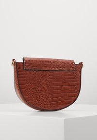 Borsa a tracolla semicerchio marrone con texture coccodrillo, chiusura con patta e anelli color oro su ogni lato, esposta su un piedistallo bianco.