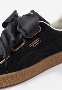 Černé sametové tenisky s tlustou hnědou gumovou podrážkou, zlatými akcenty a velkými černými saténovými tkaničkami ve tvaru mašle. Obsahuje značení "PUMA BASKET".