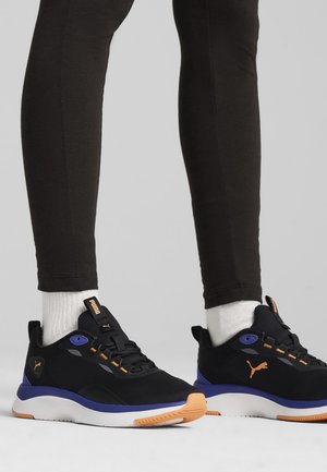 Persona con zapatillas Puma negras, azules y naranjas, con calcetines blancos y leggins negros, de pie sobre un fondo gris.