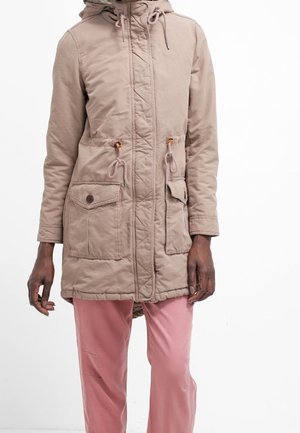 Personne portant un parka à capuche beige avec des poches avant et un pantalon rose, debout devant un fond blanc.