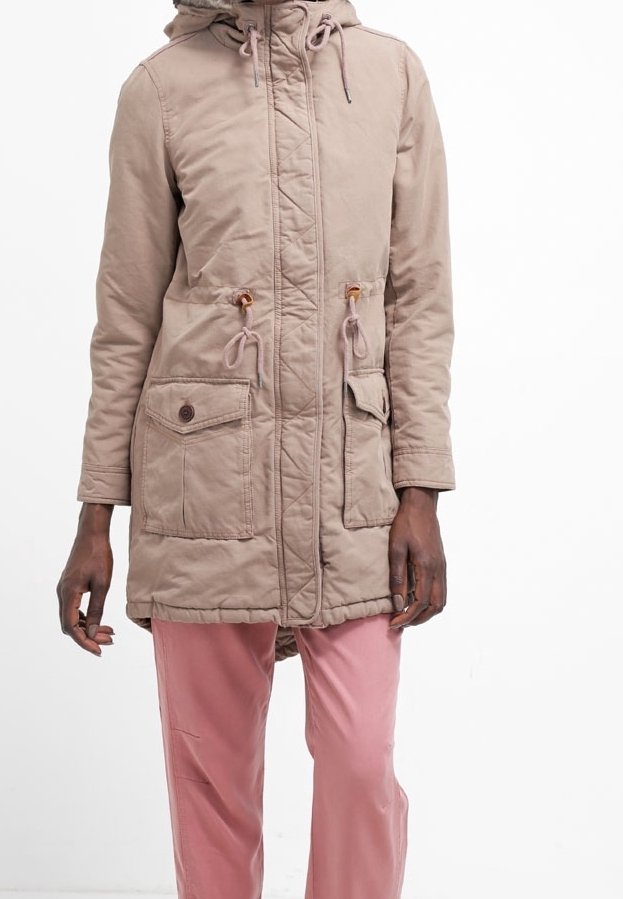 Personne portant un parka à capuche beige avec des poches avant et un pantalon rose, debout devant un fond blanc.