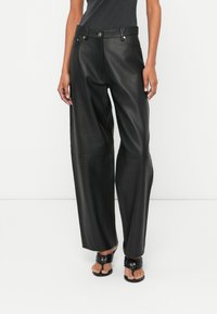 Vrouw in hoge taille zwarte leren wijde broek met een voorpaneel met knoop en rits, gecombineerd met zwarte open-toe hakken.