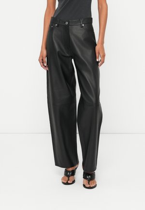 Femme portant un pantalon en cuir noir taille haute à jambes larges avec bouton et fermeture éclair à l'avant, associé à des sandales à talons ouverts noires.