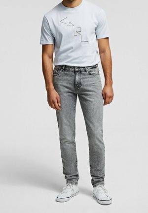 Homme portant un t-shirt bleu clair avec le logo KARL, un jean gris délavé et des baskets blanches, debout devant un fond uni.