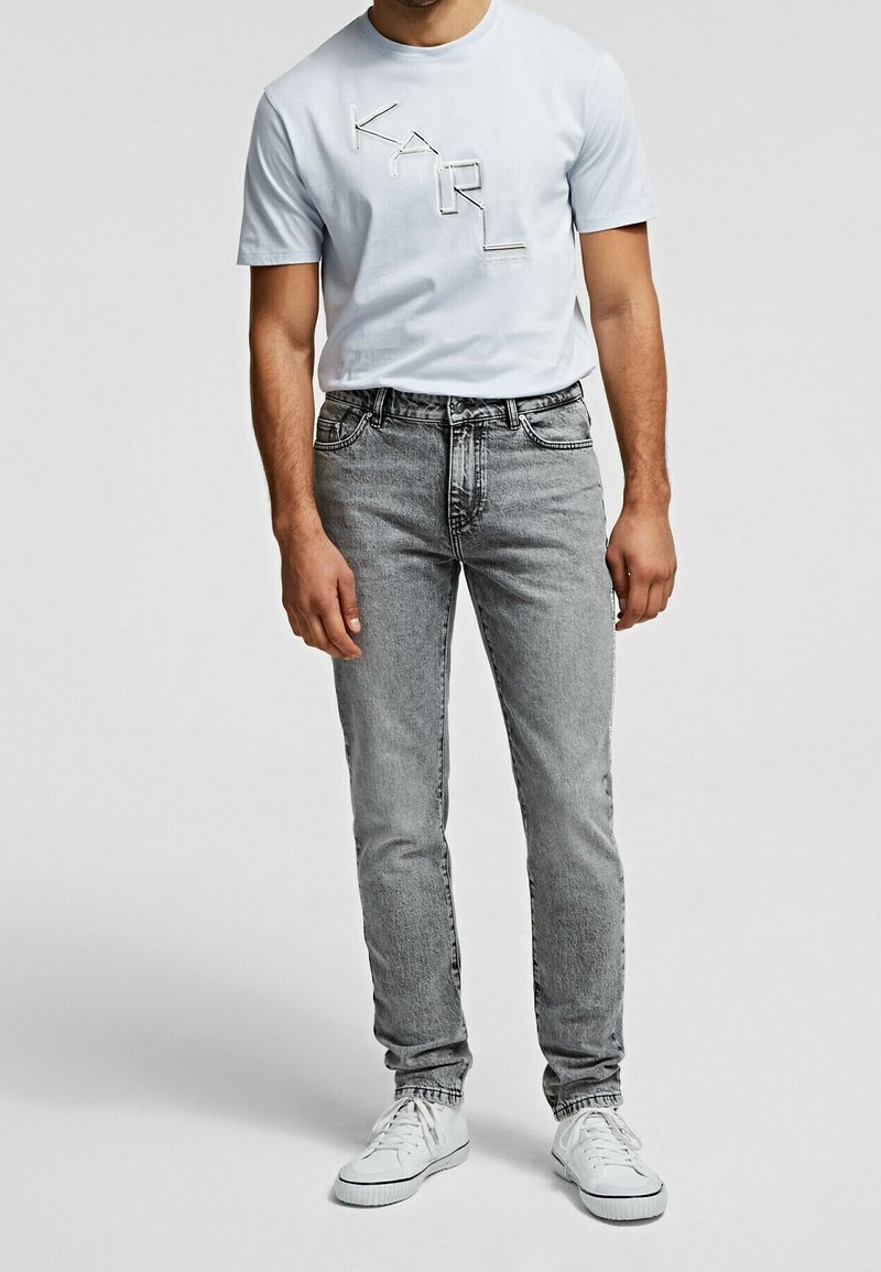 Homme portant un t-shirt bleu clair avec le logo KARL, un jean gris délavé et des baskets blanches, debout devant un fond uni.