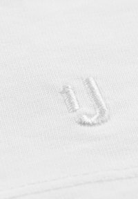 Vit textil med ett strukturerat stickat mönster, med en liten vit broderad "U" detalj synlig i förgrunden.