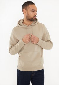 Beżowa bluza z kapturem, z kangurzą kieszenią, ściągaczami przy rękawach i u dołu. Miękki materiał o gładkiej teksturze, noszona na ciemnych dżinsach.