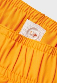 Cinturilla elástica naranja con textura fruncida, con una etiqueta de malla blanca y un cierre de botón translúcido.