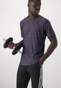 Maglietta sportiva a righe viola con maniche corte e un logo nero, abbinata a pantaloni neri con strisce bianche ai lati. Tiene un manubrio.