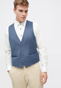 Μπλε υφασμάτινο γιλέκο με λεία υφή, διακοσμημένο με πέντε κουμπιά, βαθύ V ντεκολτέ και πλαϊνές τσέπες, συνδυασμένο με λευκό πουκάμισο.