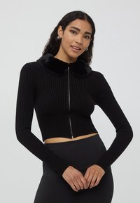 Czarny krótki kardigan z ribbowej tkaniny z długimi rękawami i pluszowym czarnym kołnierzem z sztucznego futra. Posiada srebrny zamek błyskawiczny.