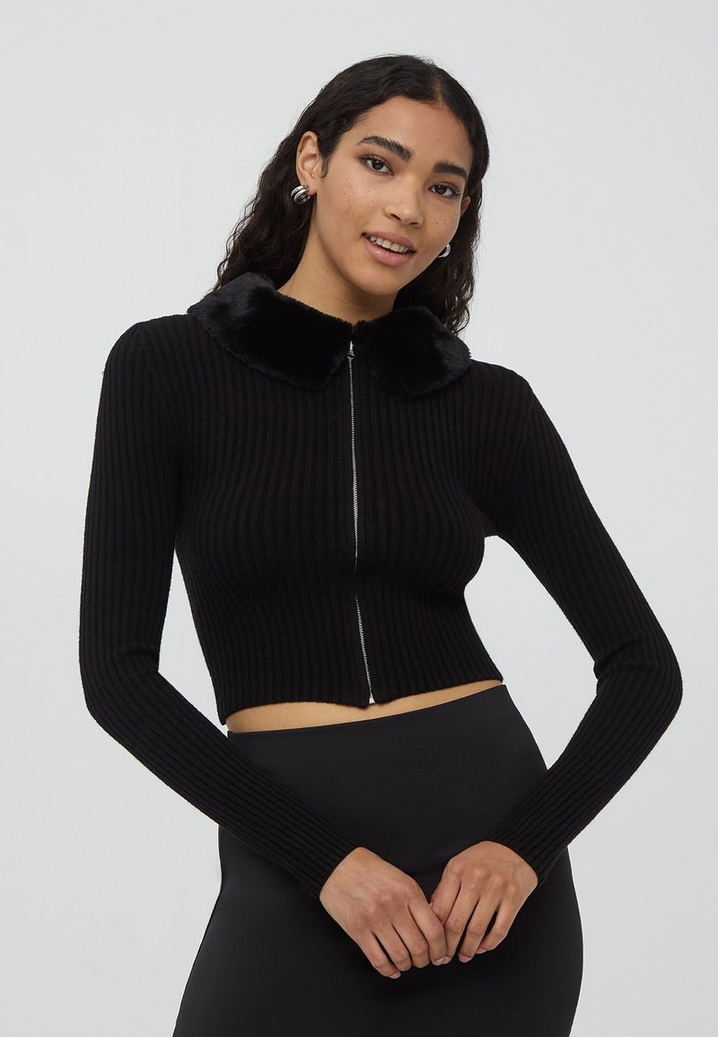 Czarny krótki kardigan z ribbowej tkaniny z długimi rękawami i pluszowym czarnym kołnierzem z sztucznego futra. Posiada srebrny zamek błyskawiczny.