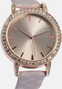 Roséguld klocka med rund urtavla, silverfärgad skiva, kristalldetaljer och ett metallic lavendelfärgat armband. Har slanka timmarkörer och visare.