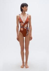 Γυναίκα που στέκεται ξυπόλητη φορώντας ολόσωμο μαγιό σε σκουριά χρώμα με αφηρημένα σχέδια και ριγέ ζώνη, κοιτάζοντας προς τα αριστερά της πάνω σε λευκό φόντο.