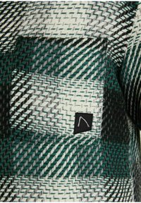 Närbild av ett vävt tyg med ett grönt, vitt och svart sicksackmönster som har en fyrkantig ficka med en liten svart etikett.