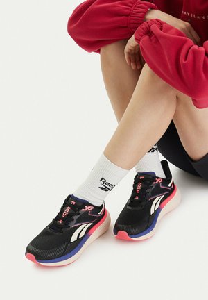 Osoba siedząca ze skrzyżowanymi nogami, ubrana w czarno-różowo-niebieskie sneakersy Reebok oraz białe skarpety Reebok, w połączeniu z czerwonym sweatshirtem.