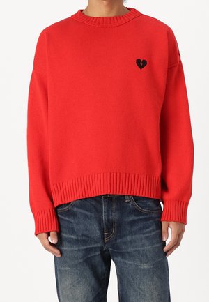 Homme portant un pull en tricot rouge vif avec un petit emblème de cœur noir brisé sur la poitrine gauche, associé à un jean en denim bleu.