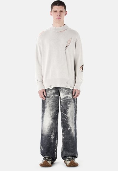 Młody mężczyzna w zużytym białym swetrze z dziurami, wyblakłych czarno-białych szerokich dżinsach oraz brązowych trampkach z jasną podeszwą, stojący prosto.