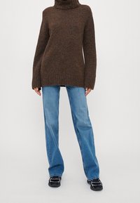 Brauner Strickrollkragenpullover mit lockerer Passform und gerippten Bündchen, kombiniert mit hellblauen, geraden Jeans und schwarzen Turnschuhen.