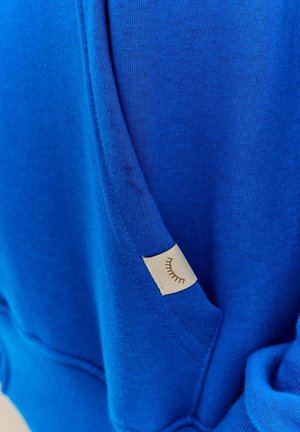 Głęboki niebieski sweter z fakturowanej tkaniny, z małą kremową metką o minimalistycznym wzorze oraz motywem rzęs na kieszeni.