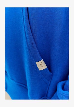 Głęboki niebieski sweter z fakturowanej tkaniny, z małą kremową metką o minimalistycznym wzorze oraz motywem rzęs na kieszeni.