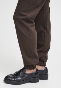 Zwarte instappers met een gladde leren afwerking en een stevige hak, gecombineerd met bruin aansluitende joggers met elastische boorden.