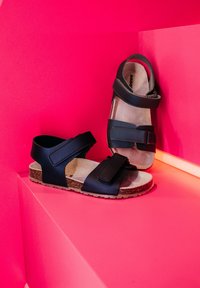 Černé sandály se dvěma nastavitelnými pásky, korkovou podrážkou a měkkou béžovou stélkou, vystavené na živém růžovém pozadí.