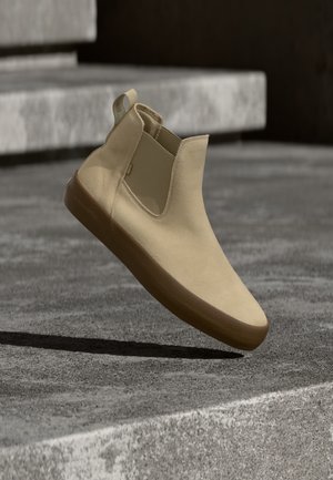 Beige slip-on ruskindsko med brunt gummisål, svævende over betontrapper i udendørs omgivelser.