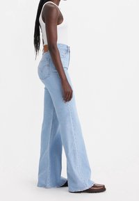 Kvinna som bär en vit ärmlös topp, ljusblå högmidjade vida jeans och bruna platåskor, står i profil.