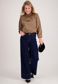 Bruine blouse met patroon en een gesmockte hals, gecombineerd met wijde donker denim jeans met zichtbare naden en een kleine zwarte bonten clutch.