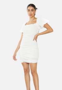 Mujer posando con un vestido corto blanco con fruncidos y mangas abullonadas sobre un fondo claro y liso.