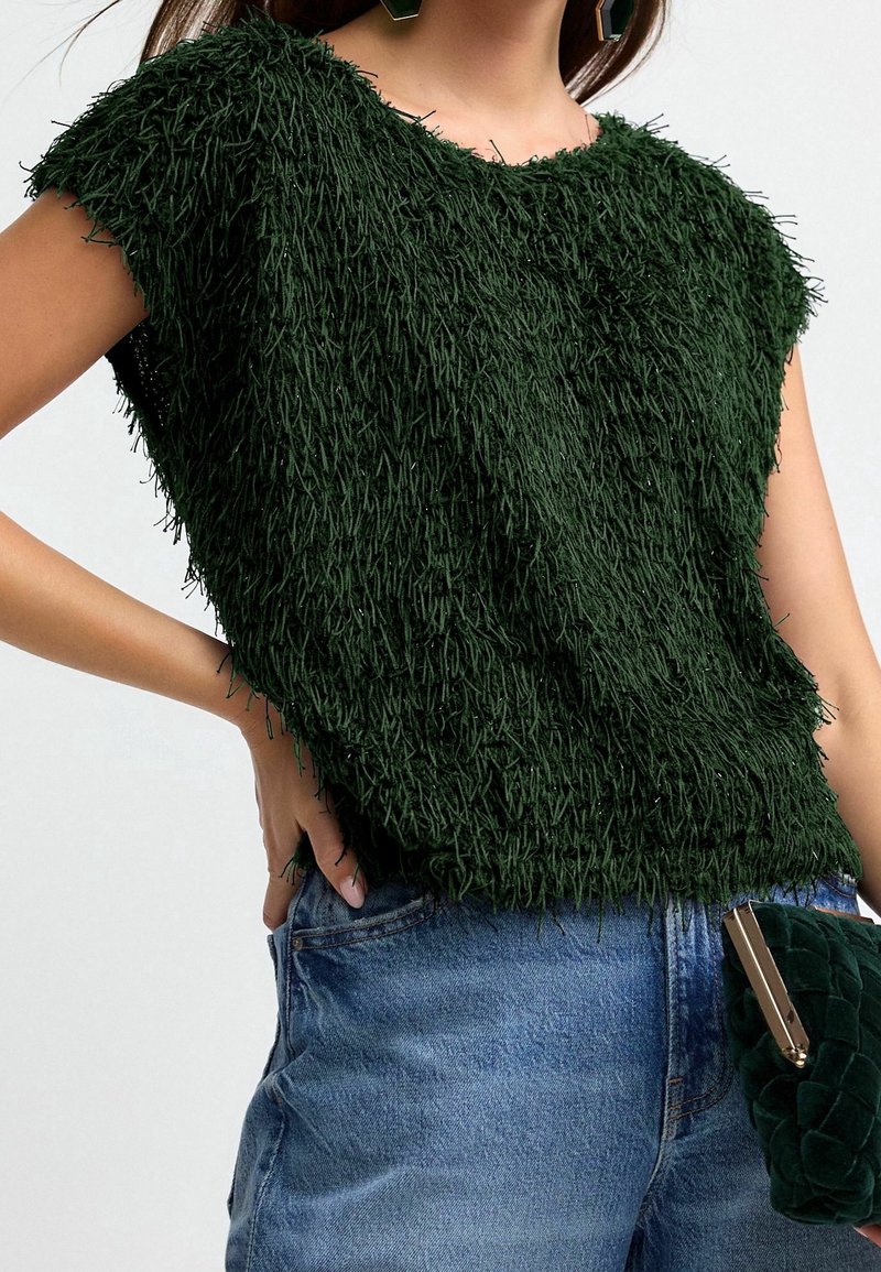 Femme portant un haut texturé à manches courtes vert foncé, un jean bleu et tenant une pochette matelassée vert foncé.