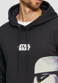 Czarna bluza z kapturem z białym logo "STAR WARS" i grafiką szturmowca. Teksturowany materiał z regulowanymi sznurkami.