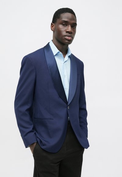 Námornícky blejzer s hodvábnym šálovým golierom, zapínaním na jeden gombík a bočnými vreckami, skombinovaný so svetlomodrou košeľou a tmavými nohavicami.