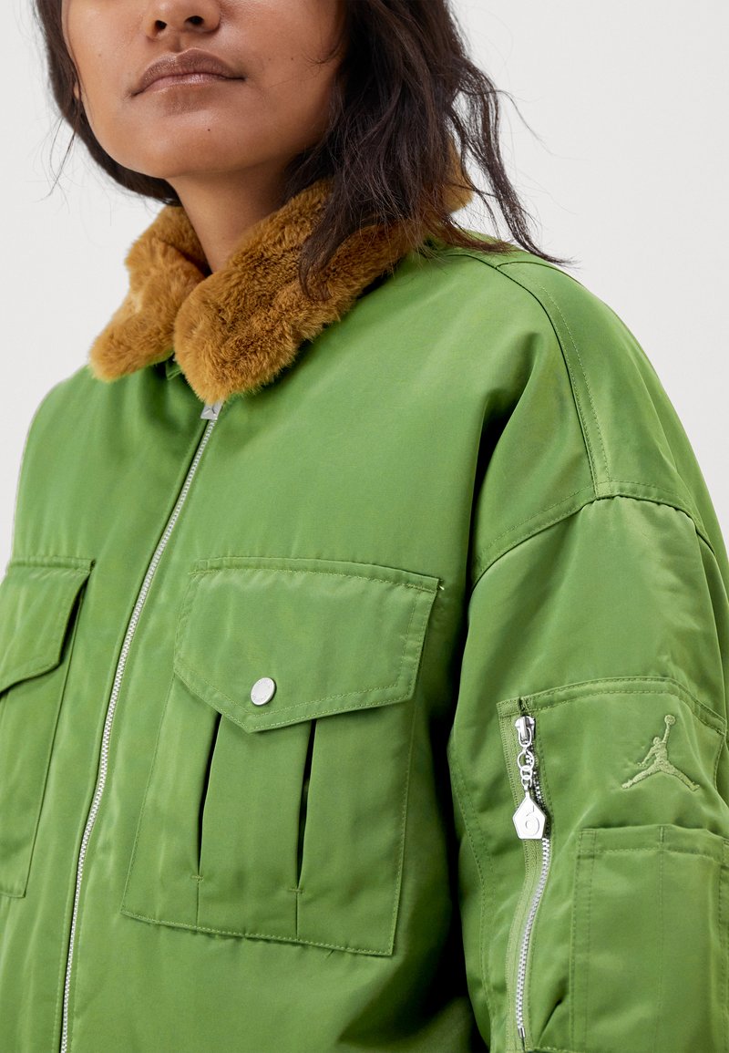 Giacca bomber verde con collo in pelliccia sintetica, tasche con zip e tasche frontali con patta. Presenta una chiusura con zip e il logo Jordan sulla manica.