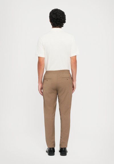 Lysfarvet polo shirt til mænd og brune bukser med elastisk talje og to baglommer, iført med sorte sko. Synlig glat stofstruktur.