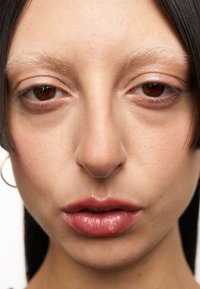 Gros plan sur un visage avec une peau lisse et claire, des yeux marron et des lèvres roses brillantes ; traits comprenant un nez droit, des sourcils clairs et clairsemés, et des cheveux noirs.