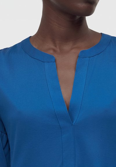 Κοντινό πλάνο ατόμου που φορά μπλε μπλουζάκι με V-λαιμόκοψη και στρογγυλεμένο γιακά, δείχνοντας το πάνω μέρος του στήθους και το λαιμό πάνω σε απλό φόντο.