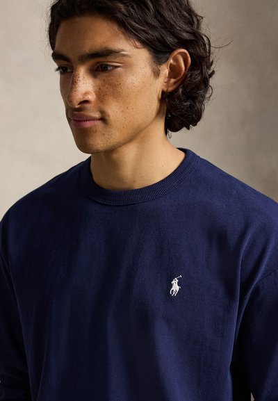 Νέος μεσαίου μήκους σκούρα μαλλιά και φακίδες που φοράει ένα μπλε σκούρο πουλόβερ με ένα μικρό λευκό λογότυπο παίκτη πόλο.