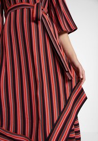 Robe rayée présentant des bandes verticales rouges, noires et blanches, avec une ceinture à la taille et un tissu fluide qui tombe avec un ourlet contrastant.