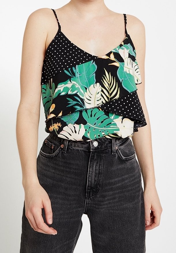 Femme portant un haut noir sans manches avec des motifs de feuilles tropicales et de pois, associé à un jean noir taille haute.