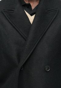 Cappotto nero in misto lana con design doppiopetto, rever a lancia e due bottoni neri, caratterizzato da una texture morbida e una forma strutturata.