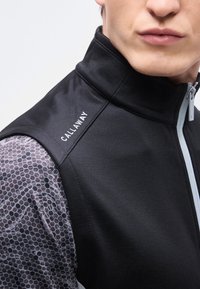 Άνδρας που φοράει μαύρο γιλέκο Callaway με γκρι πουκάμισο μακρυμάνικο με σχέδιο κυψελοειδές, εμφανίζοντας λεπτομέρειες του φερμουάρ και του γιακά.