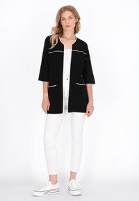Schwarze Strickjacke mit weißer Paspelierung, offene Vorderseite und Dreiviertelärmel. Wird über einem weißen Oberteil getragen und mit weißen Hosen und Sneakers kombiniert.