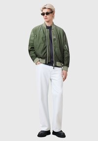 Olive green bomber jacket with side pockets, paired with a navy shirt, white wide-leg pants, and black shoes. Model wears dark sunglasses.