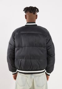 Black puffer jacket with quilted texture, ribbed black and white cuffs, and a rounded collar. Styled with light blue jeans.