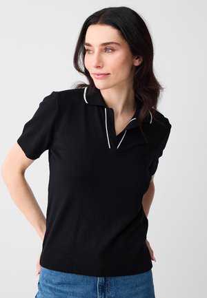 Frau mit dunklem Haar, die ein schwarzes Poloshirt mit weißen Akzenten und kurze Ärmel sowie eine blaue Jeans trägt, schaut leicht nach rechts und hat die Hände hinter dem Rücken.