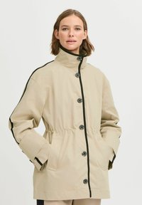 Beige sako na knoflíky s černými akcenty, vysokým límcem, stahovacími pasem a dlouhými rukávy; vyrobeno z lehkého materiálu.