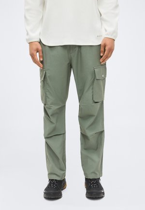 Homme portant un pantalon cargo vert olive avec des poches latérales, des chaussures de randonnée noires et une chemise blanche à manches longues, debout devant un fond uni.